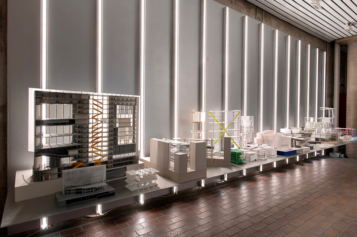 A variety of architectural models on a metal display shelf with vertical lighting behind them.