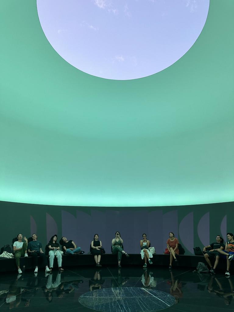 People seated on a bench in a curved room with a green ceiling. In the center is a circular window.