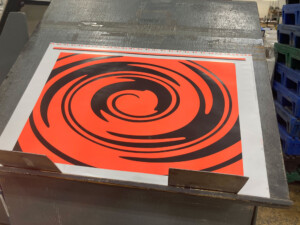 The GSD public programs poster in the process of being printed. The black-and-red spiral on the poster sits on a metal table.