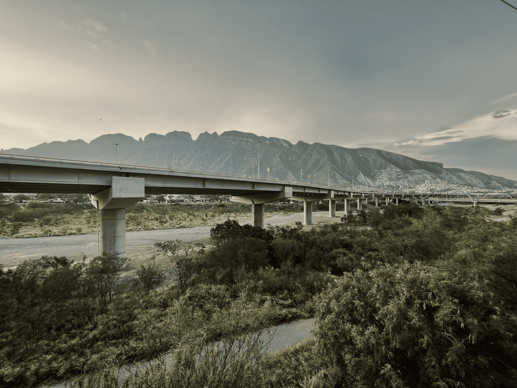 A highway rises over a dry riverbed filled with trees. Mountains are in the background.