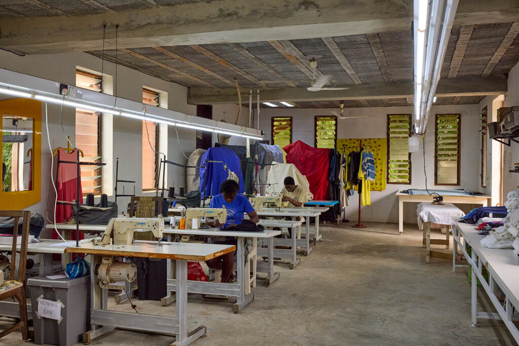 Photo of large room set up as a working space with sewing machines on the tables. Two people are sewing. The room has open windows all around.