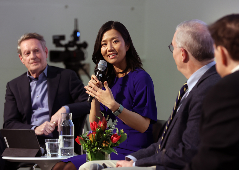 Mayor Michelle Wu holds a microphone and appears to be speaking. She is seated among a group that includes three men. On a small table in front of her are water glasses, a pitcher, and an ipad device.
