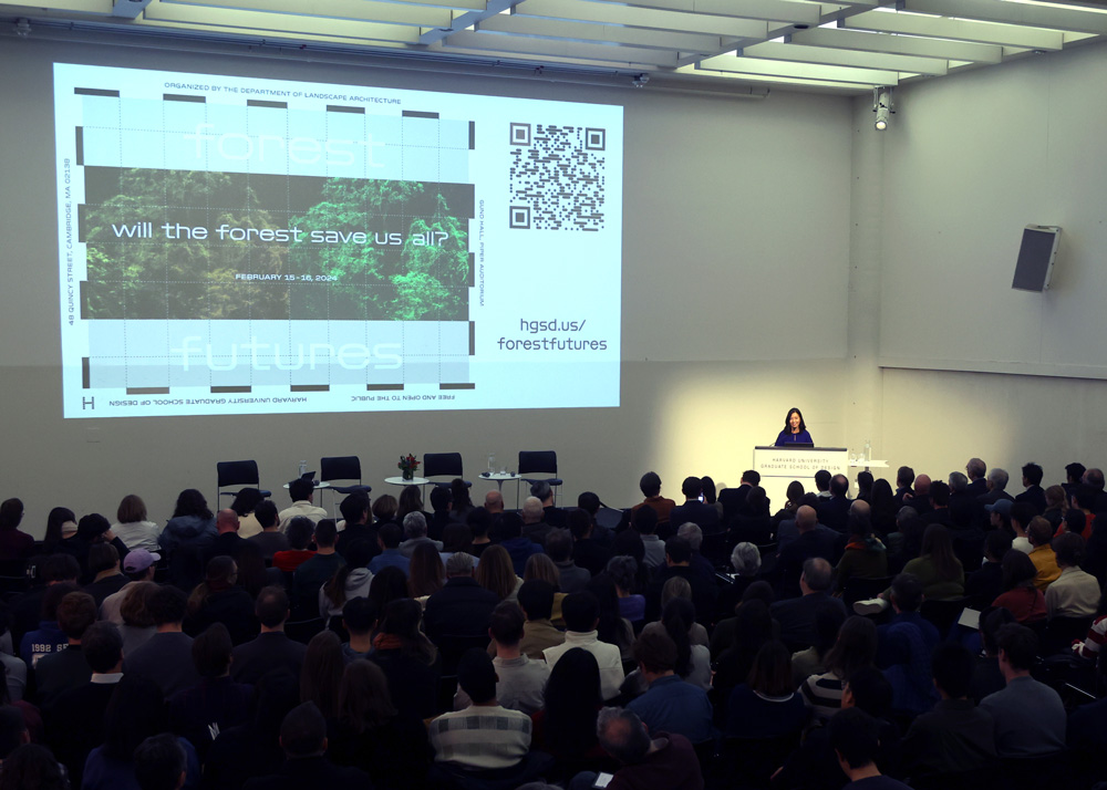 Mayor Michelle Wu stands at a podium in the Harvard Graduate School of Design's Piper Auditorium. An audience of more than fifty people is visible. A slide projected on a nearby screen says: "Will the forest save us all?"