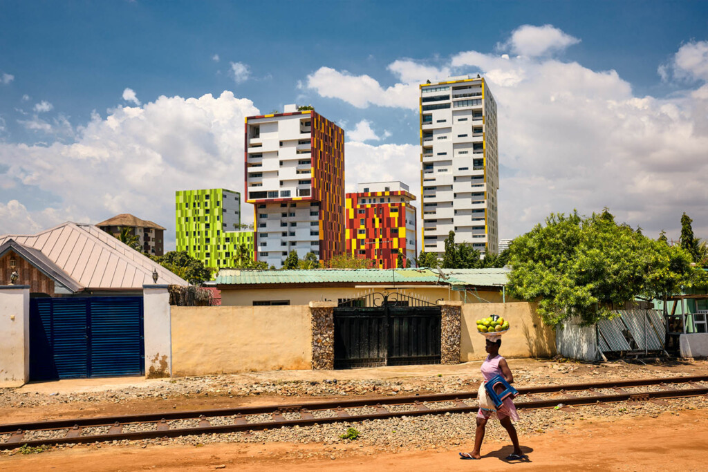 African woman walking with a basket of fruit on her head in front of four colorful tall buildings. The railroad is in front of her.
