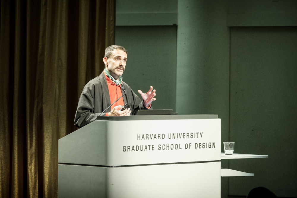 Daniel Fernandez Pascual stands at a podium on which appears the text Harvard University Graduate School of Design. He wears a black coat over a red sweater. He appears to be speaking to the audience.