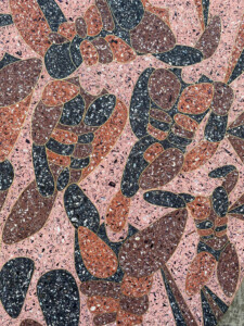 An abstract pattern in pink, black, brown, and light red that resembles a group of body builders posing to display their muscles. The material is flecked with white and black shiny pieces of mussel shell.
