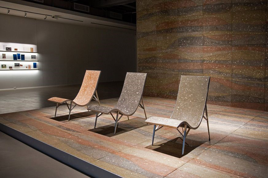 Three modernist lounge chairs site on a platform adorned with a wavy abstract pattern in dull colors. The surfaces of the chairs and platform are flecked with white.