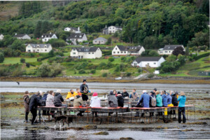 A group of people sit at an installation of tables and benches made from metal oyster cages. The installation sits in a muddy intertidal zone. 