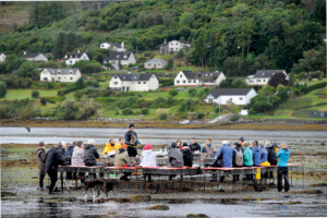 A group of people sit at an installation of tables and benches made from metal oyster cages. The installation sits in a muddy intertidal zone.