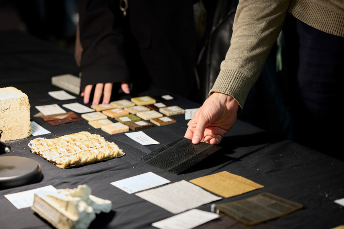 Samples of different materials made of organic waste and fibers are arrayed on a black cloth on a table. Two hands appear in the image touching different materials.