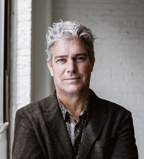 A portrait of John Ronan, a man with grey hair who wears a dark blazer and a dark shirt with light spots on it.