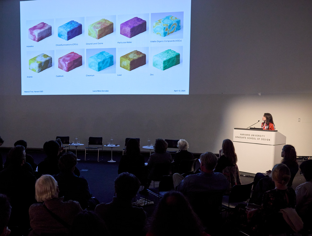 A woman in a red shirt with long dark hair speaks at a podium at the Harvard Graduate School of Design. On the screen behind her and to her right is a larger projected image of bricks in many different colors and patterns.
