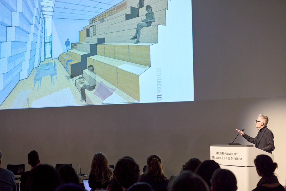 A man in black clothing and glasses with black frames speaks at a podium at the Harvard Graduate School of Design. Behind him and to his right is a large projected digital image of an interior space with large steps toward one side on which people sit and read.