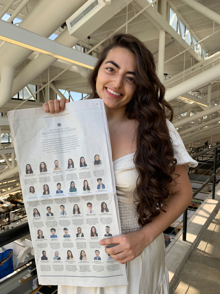 A woman stands in the studio area of Gund Hall holding a newspaper to a page that features thumbnail portraits of different people.