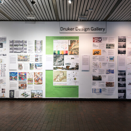 A view of the exhibit in Gund Hall, showing a variety of student work hanging on the walls.