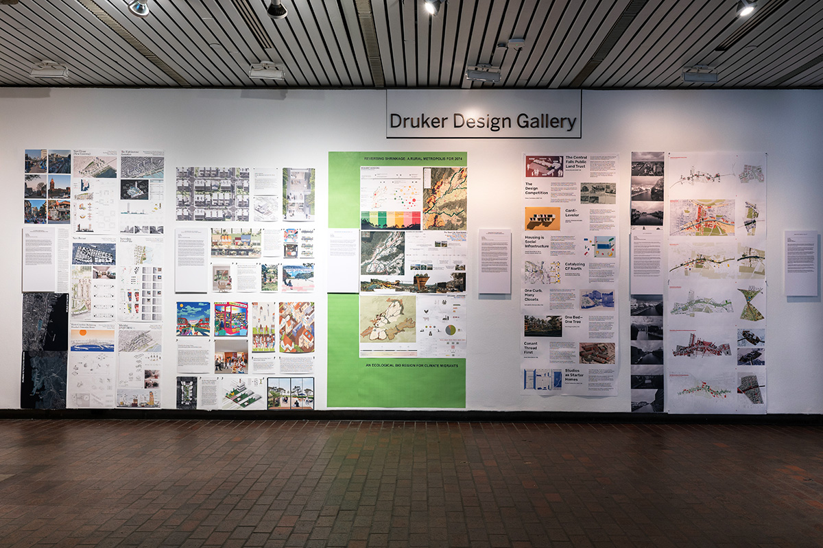 A view of the exhibit in Gund Hall, showing a variety of student work hanging on the walls.