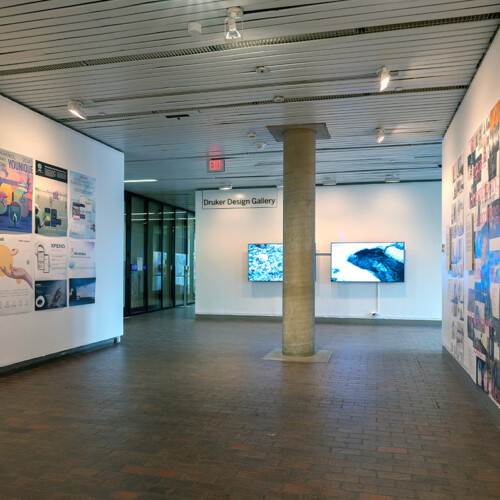 A view of the exhibit in Gund Hall, showing a variety of student work hanging on the walls.