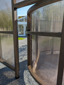 A detail view of a public artwork made of wood beams and plastic sheets. An entrance is formed as one curved wall intersects with one straight one.