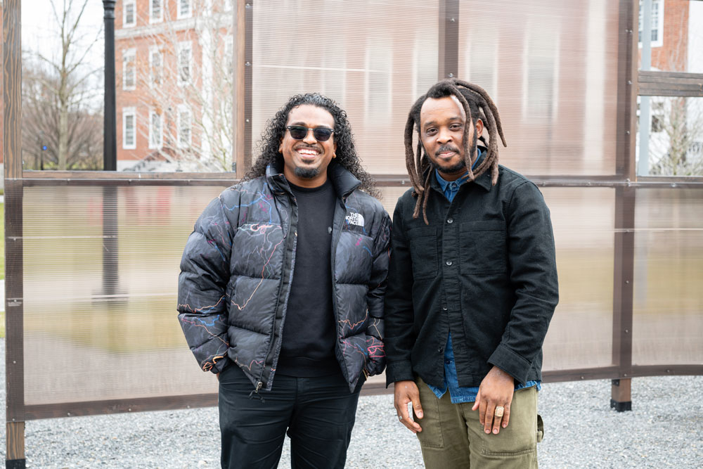 Two men stand in front of a curving wall made of wood and plastic.