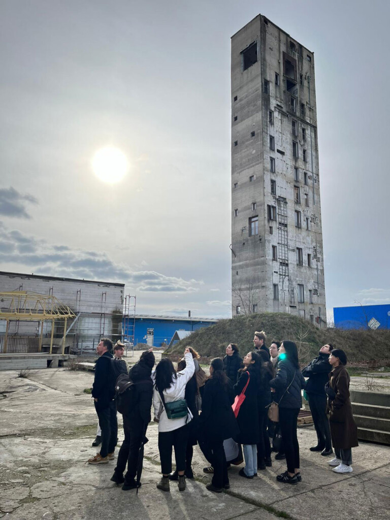 A group of fifteen people stands in an open area paved in concrete surrounded by industrial buildings in grey and blue. A tall, narrow concrete tower with irregular buildings and some apparent smoke damage stands in the open area as well.