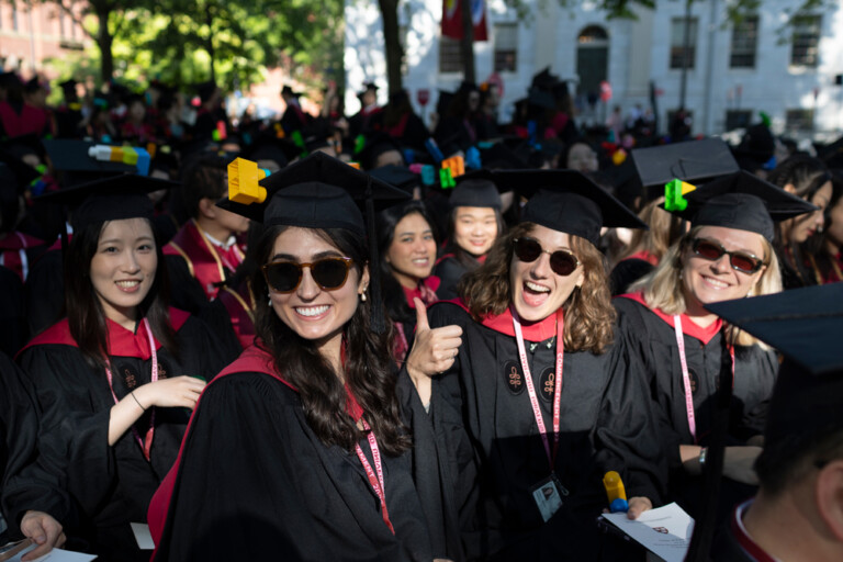 A group of people in black graduation caps and robes with red collars smile at the camera. Some have large plastic toy blocks affixed to the flat brims of their caps.