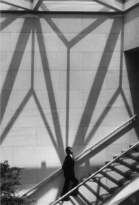 A photograph of I.M. Pei walking up a staircase in the National Gallery in Washington, D.C. Shadows defining large triangular forms are on the wall behind him.
