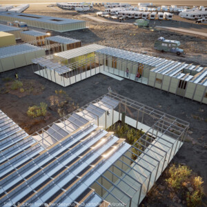 A digital rendering of a desert landscape with beige, modular structures in the foreground and a parking lot with mobile campers in the background.