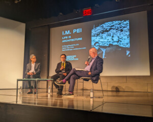 Three men sit on chairs on a stage and hold microphones. Behind them is a screen displaying a picture of I.M. Pei and the text I.M. Pei Life is Architecture.