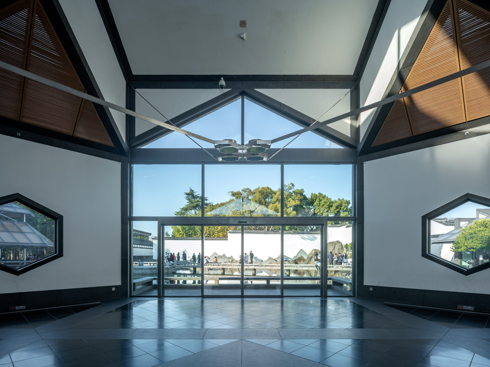 The interior of the Suzhou Museum showing an open space with two hexagonal windows and a large glass entryway looking out onto a courtyard with large rocks and a pond.