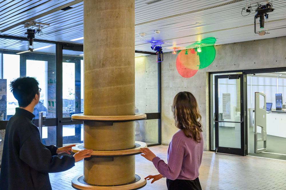 A color image of two people touching wood rings attached to a concrete column in Gund Hall at the GSD. Projectors attached to the ceiling cast colored lights on the building.