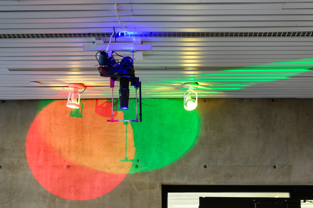 A photograph of a projector attached to the ceiling of Gund Hall. Red and green lights are cast on the wall behind it.