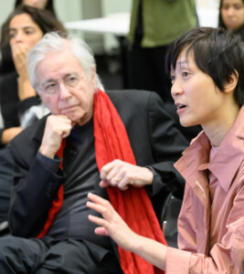 A photograph showing a woman speaking while two men listen in. Other members of the audience are in the background.