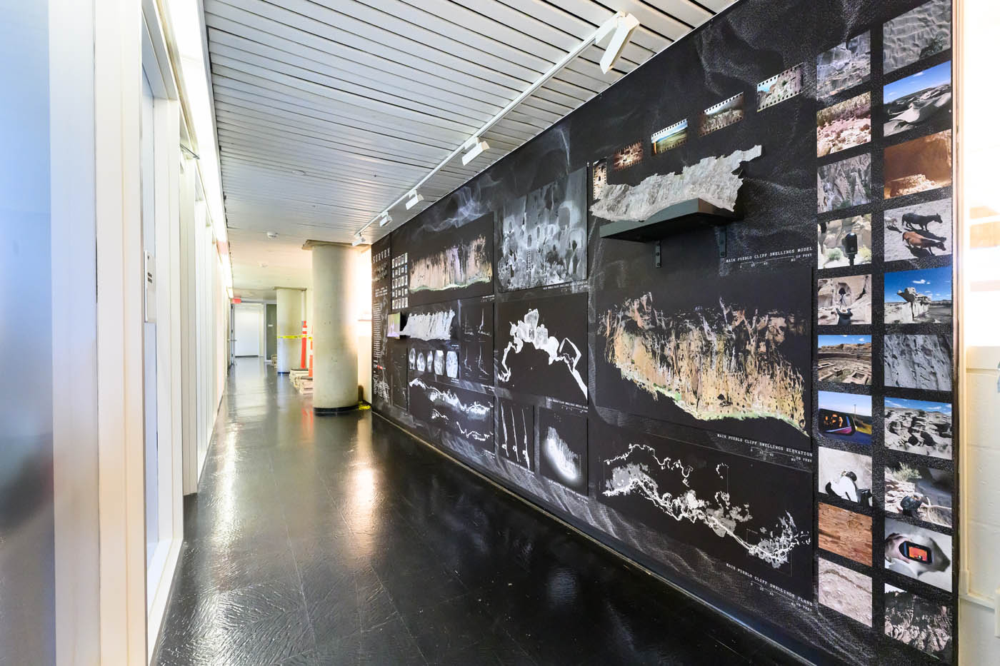 a long view down the exhibition wall, showing photographs, a video, and models