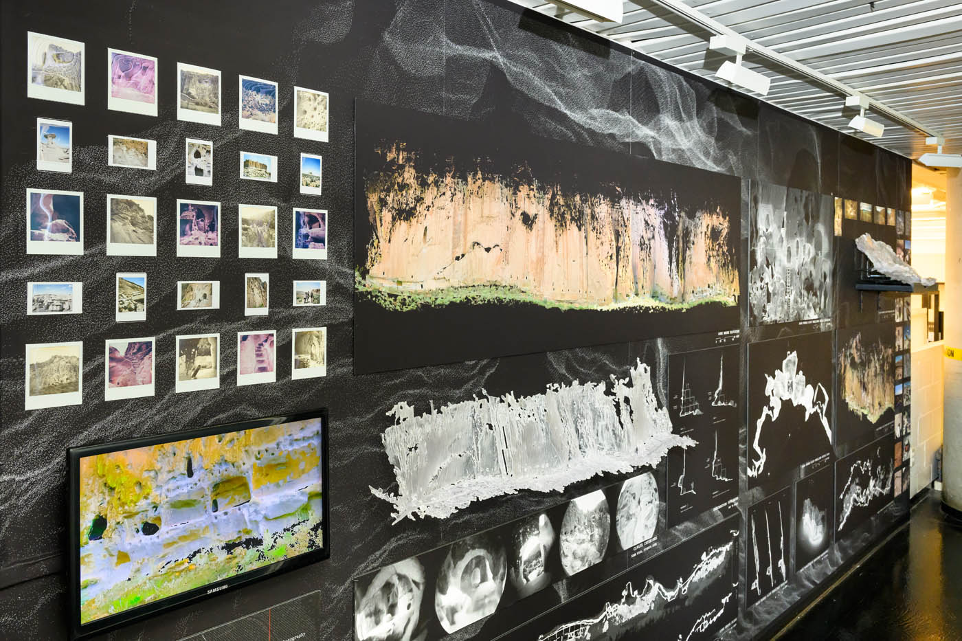 a long view down the exhibition wall, showing photographs, a video, and models