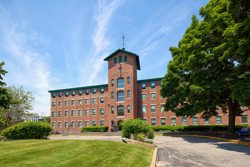 Red brick building with a lawn in front of it and a tree to the right.