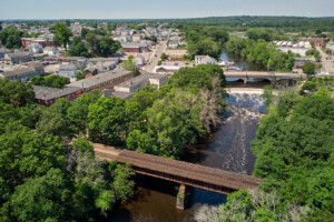 Aerial photo showing a river with two bridges. Green trees on each side of the river in the foreground and houses in the background