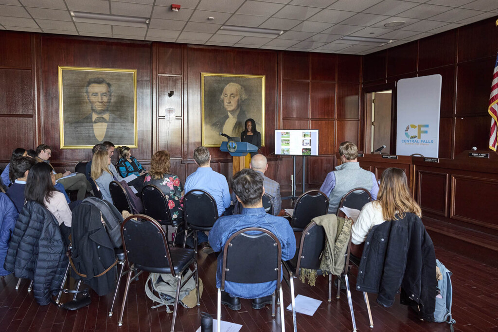 A group of people sitting in a wood paneled room in Central Falls, RI City Hall. African American student presents at the podium. Images of Abraham Lincoln and George Washington adorn the walls