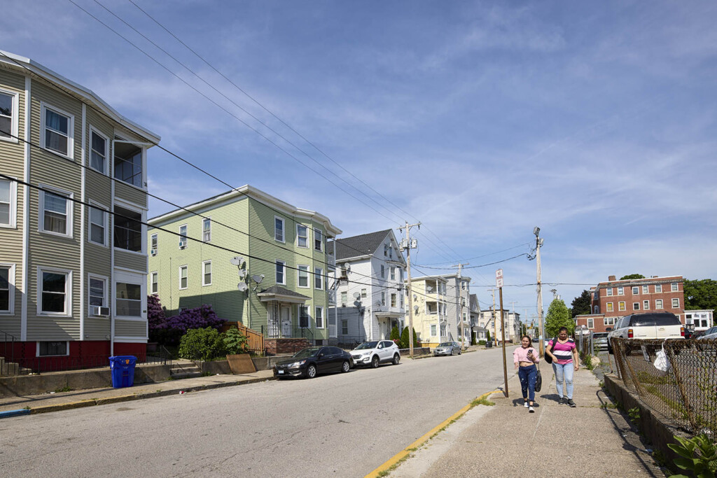 A street with row of triple decker houses on one side and two females walking on the other side of the street in Central Falls, RI