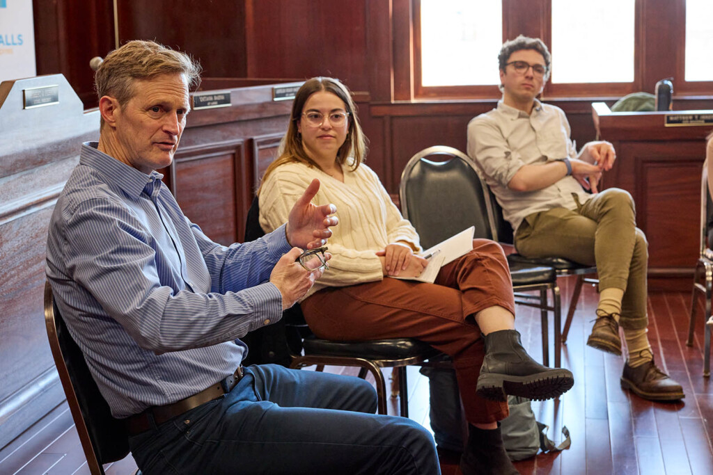 Caucasian looking male speaks gesticulating with his hands wile two students from the GSD are listening