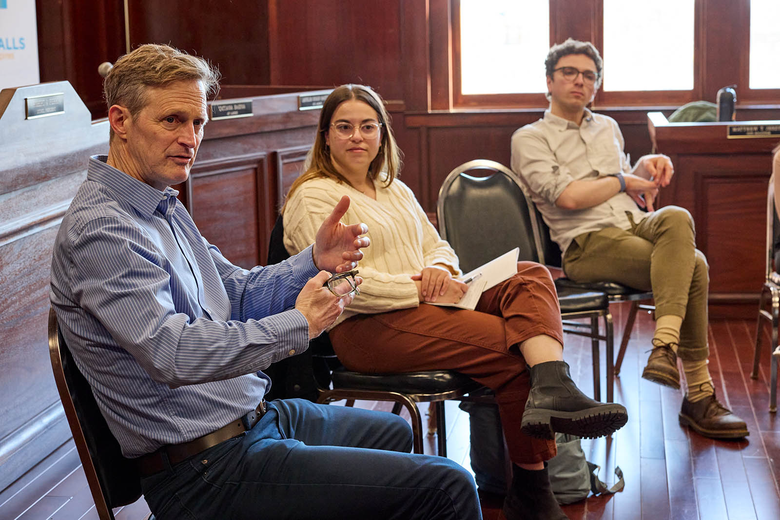 Caucasian looking male speaks gesticulating with his hands wile two students from the GSD are listening