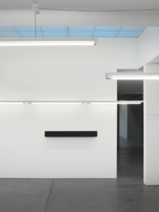 An installation view of an artwork by Sarah Oppenheimer showing a black rectangular box affixed to a white wall and white fluorescent lights overhead.