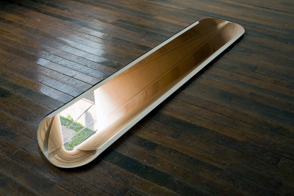 A photograph of an art installation featuring a oval hole cut into a wood floor. The hole reveals a view of an outside space on the exterior of the building.