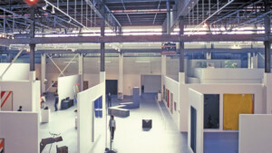 An interior view of the Geffen Contemporary at MOCA showing an installation of minimal sculptures and abstract paintings in a large, open warehouse space with temporary walls erected inside.
