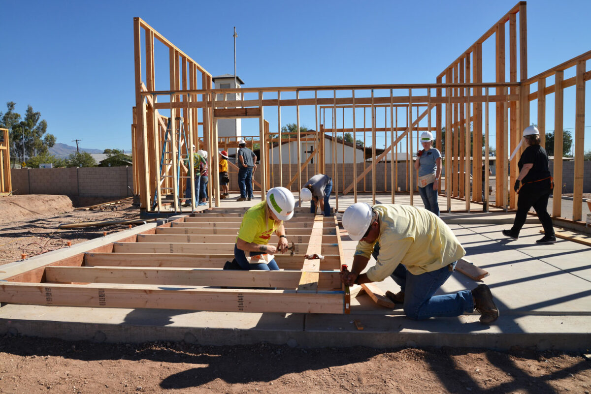 A group of people work together on a house under construction. Wood framing of the house is visible, and two people in the foreground use hammers to assemble more wood beams.