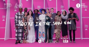 Cast of actors stand against a pink backdrop with text "You're in the Sun Now" overlayed.