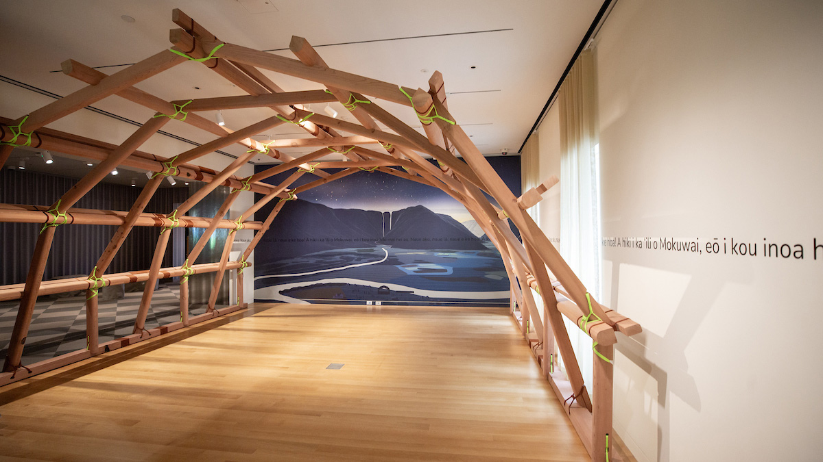 A photo showing a rounded arch structure made of wood beams held together with yellow cords. The structure is in a museum gallery whose walls are adorned with a landscape painting of mountains and a river and Hawaiian text.