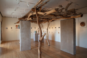 An installation in a museum gallery showing a structure made of unfinished wood logs and concrete supports.