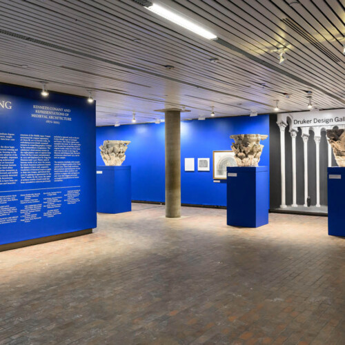 view of gallery with introductory wall and two plaster casts of column capitals on pedestals infront of images on a wall and a mural of columns.