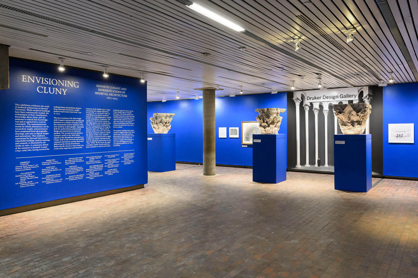 view of gallery with introductory wall and two plaster casts of column capitals on pedestals infront of images on a wall and a mural of columns.