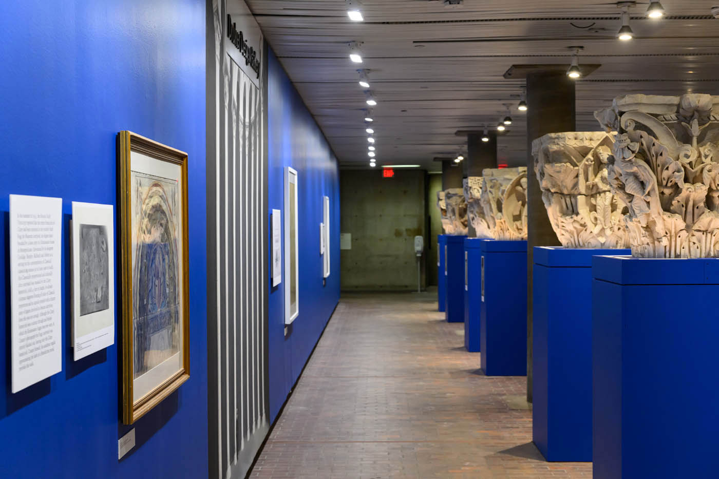 A long view of the gallery. On one side is a wall with images, framed paintings, and a mural. On the other side are four replica column capitals on pedestals.
